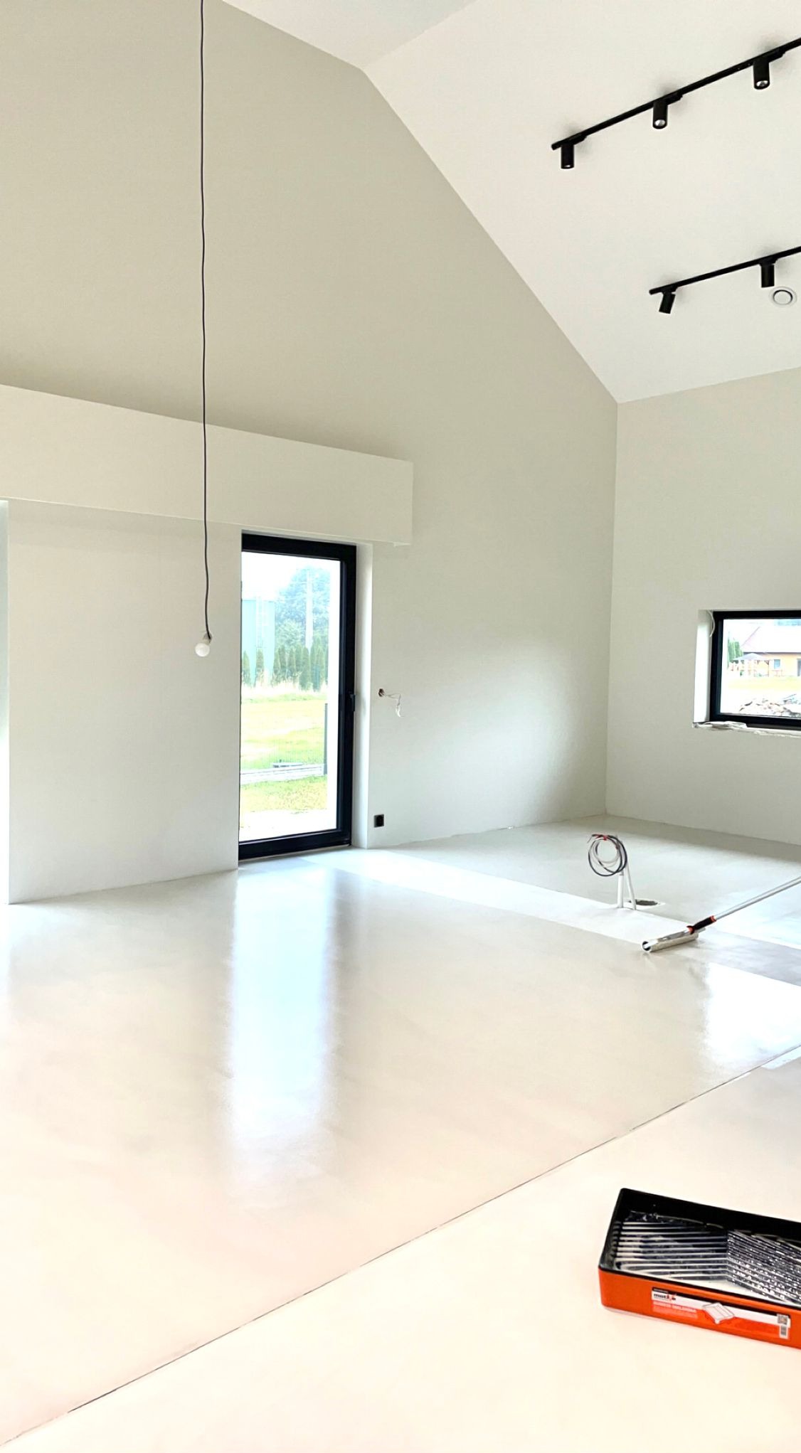 Wnętrze domu z wysokim sufitem i dużymi oknami, świeżo pomalowane na biało, z widocznym wałkiem do malowania podłogi i kuwetą z farbą w rogu