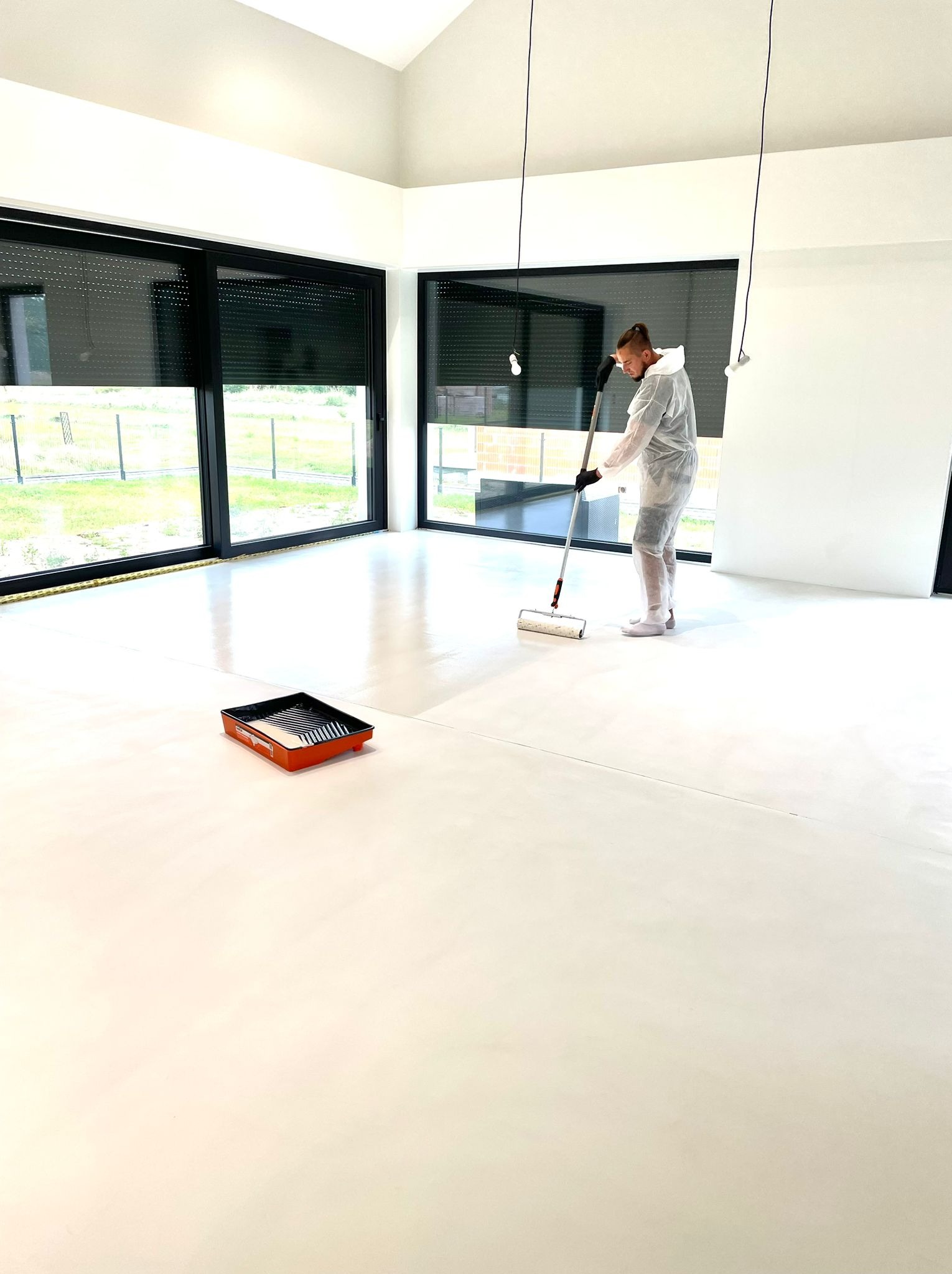 Pracownik w kombinezonie ochronnym aplikuje wałkiem jasną, błyszczącą powłokę na posadzkę w przestronnym, nowoczesnym wnętrzu z dużymi oknami i wiszącymi lampami bez kloszy. Kuweta z farbą stoi obok.
