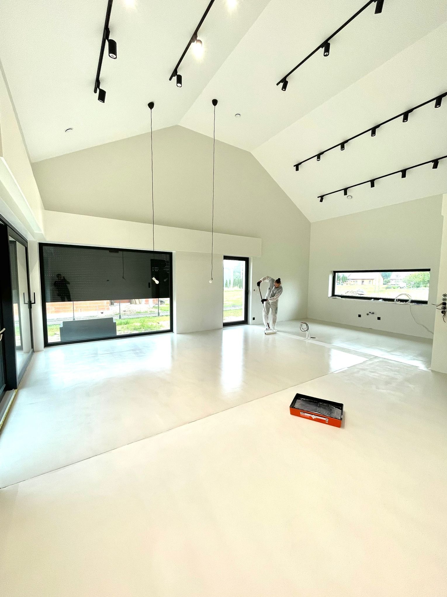 Wnętrze nowoczesnego domu z wysokim sufitem, gdzie pracownik w kombinezonie malarskim rozprowadza jasną posadzkę żywiczną za pomocą wałka, widoczna kuweta malarska na podłodze.