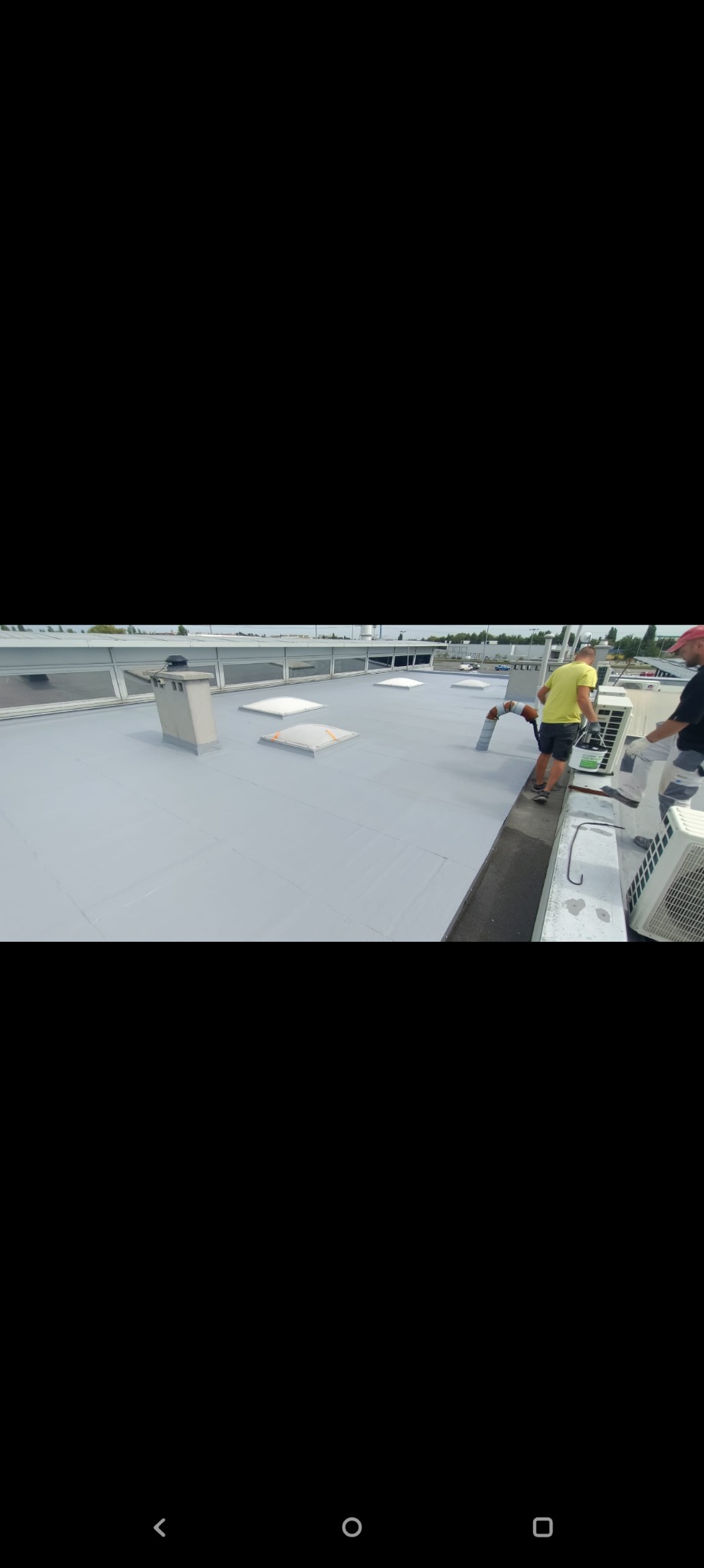 Praca na dachu: Dwóch mężczyzn w trakcie aplikacji szarej powłoki na płaskim dachu budynku, widoczne kominy wentylacyjne i klimatyzatory.