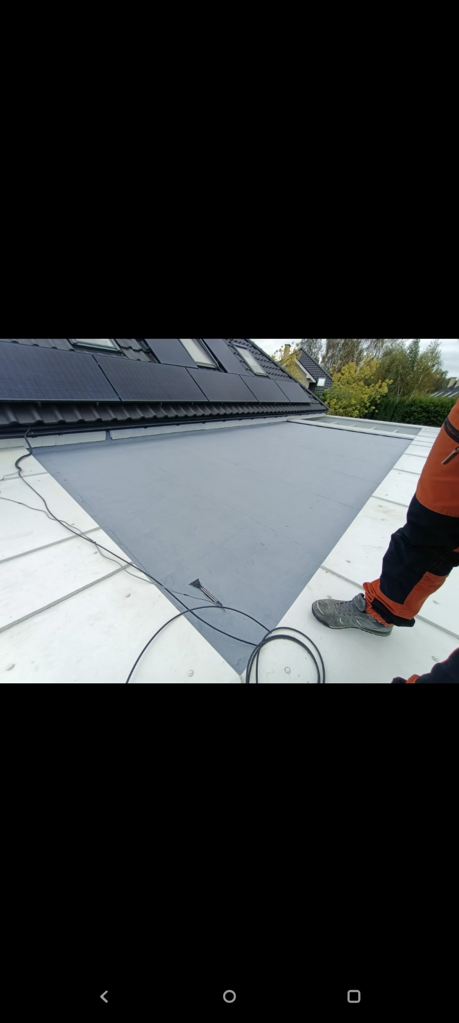 Szary dach z panelami słonecznymi i oknami dachowymi, częściowo pokryty nową powłoką. Widoczne buty i fragment stroju roboczego osoby wykonującej prace.
