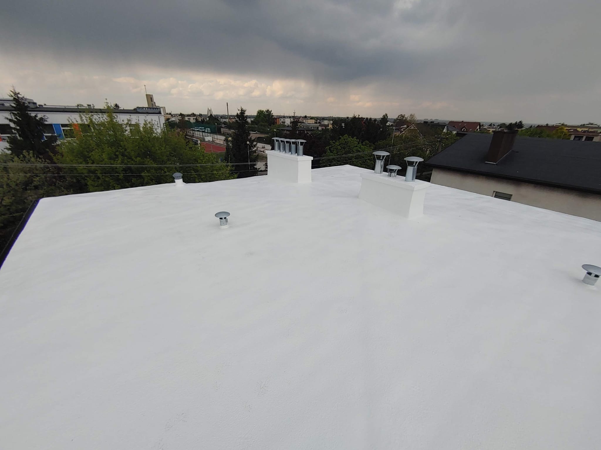 Biały, płaski dach po renowacji w Kaliszu, widok z góry. Kominy i wentylatory na tle pochmurnego nieba i zabudowań miejskich. Nowa izolacja dachu.