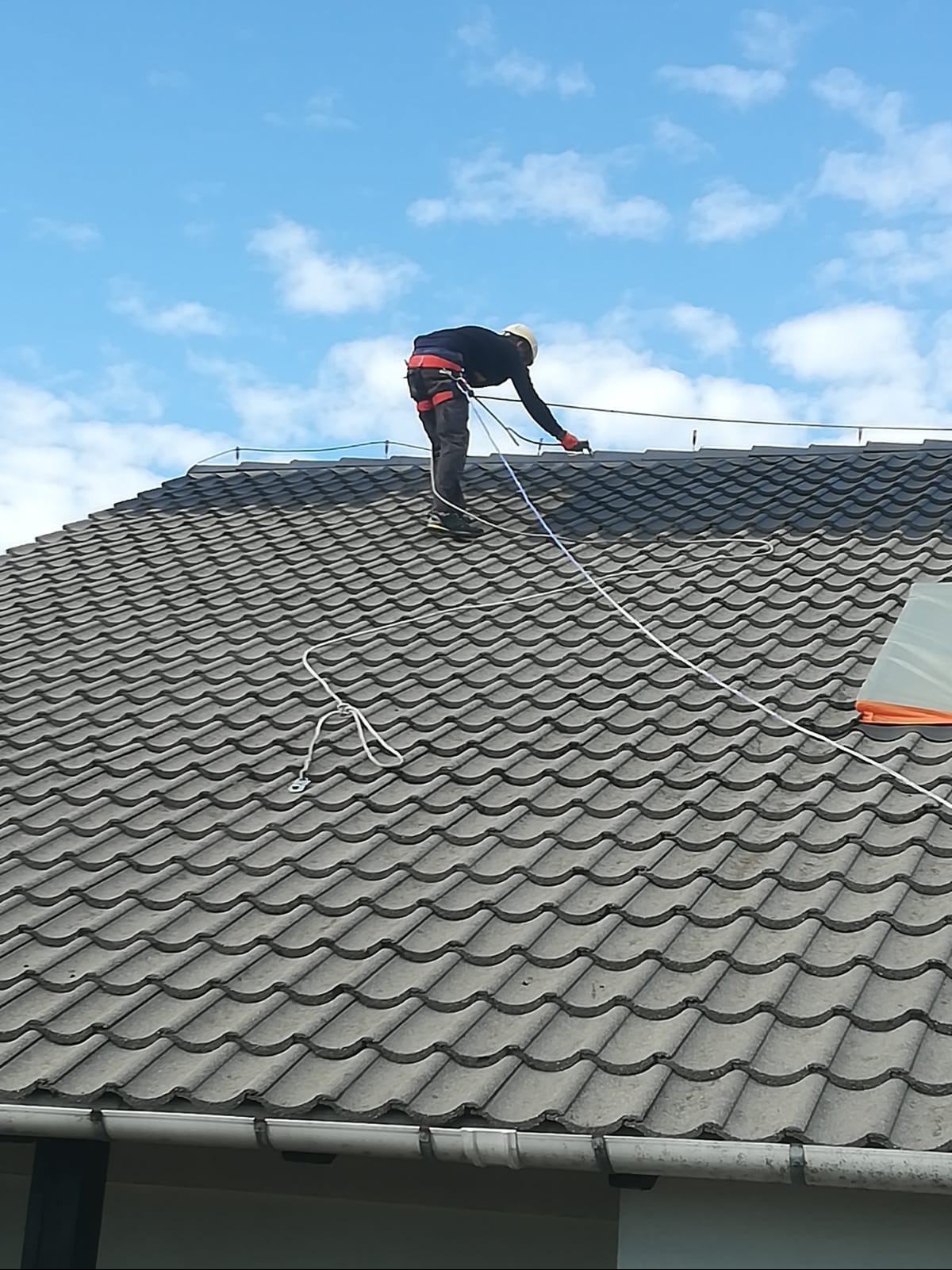 Pracownik w kasku i uprzęży zabezpieczającej na szarym dachu z dachówki ceramicznej, podczas montażu zabezpieczeń. Błękitne niebo w tle.
