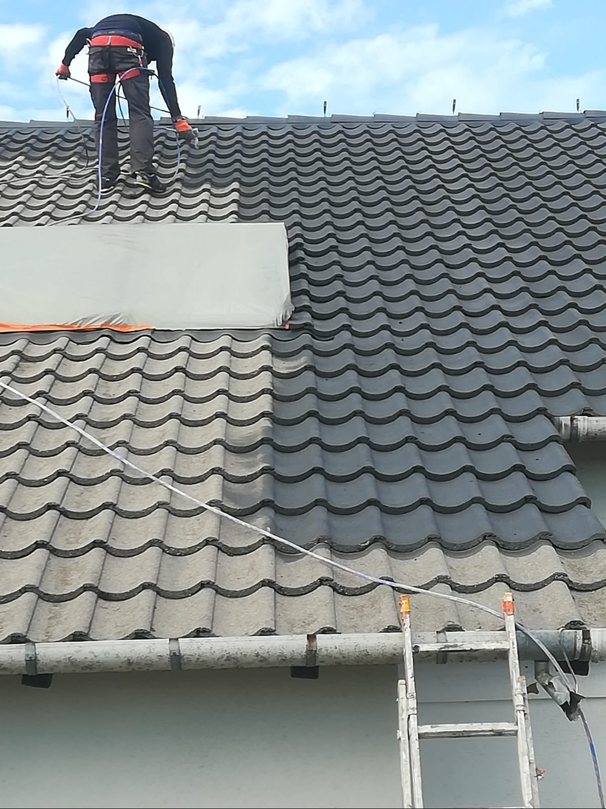 Pracownik z zabezpieczeniami maluje dachówkę natryskowo. Część dachu pomalowana na szaro, część w kolorze naturalnym. Widoczna drabina i zabezpieczone fragmenty dachu.