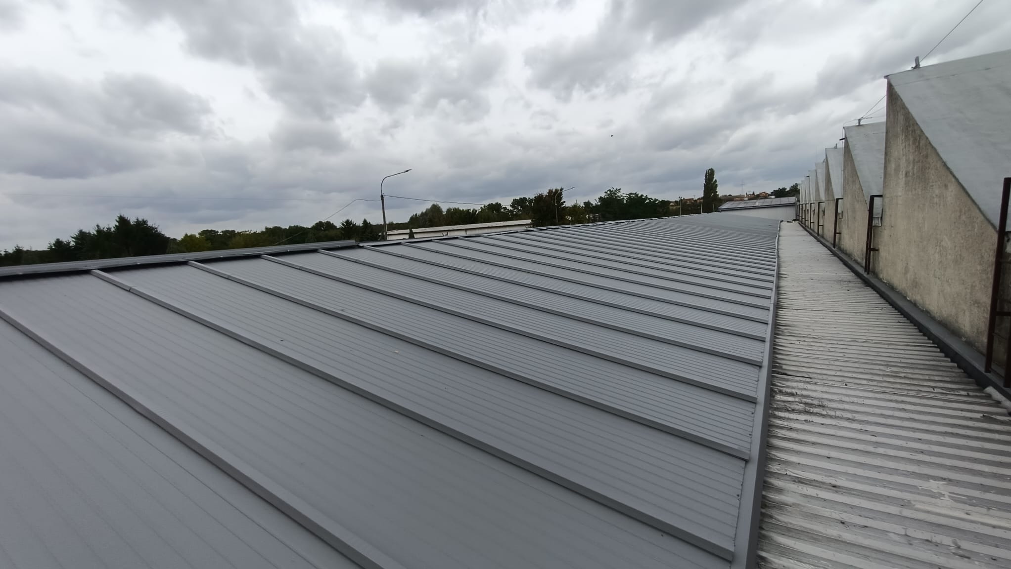 Szary dach z blachy trapezowej na budynku przemysłowym pod pochmurnym niebem. Widoczny fragment starego dachu i pionowe elementy konstrukcyjne.