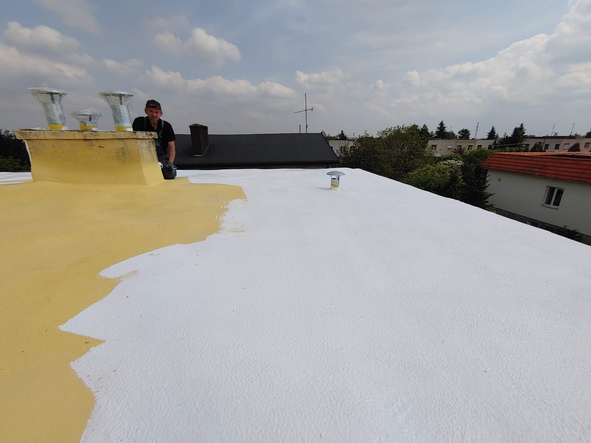 Biała powłoka na dachu w trakcie aplikacji, widoczny fragment żółtej, oryginalnej powierzchni. Pracownik w tle. Dach z kominami na tle zabudowań.