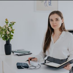 Angelika Kukulska - dietetyk kliniczny Kolbuszowa - Dietetyczka w białej koszuli, mierząca ciśnienie pacjentowi przy biurku z wazonem kwiatów, notując wyniki w otwartym notatniku.