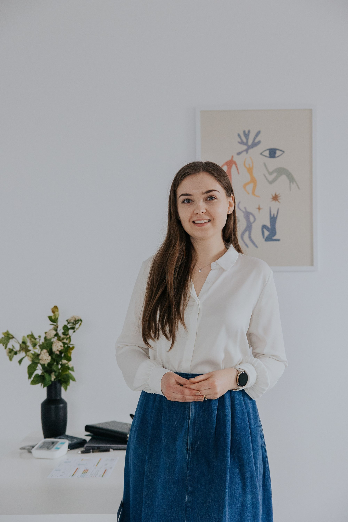 Portret uśmiechniętej kobiety w białej koszuli i dżinsowej spódnicy, stojącej w jasnym biurze z abstrakcyjnym obrazem na ścianie i wazonem z kwiatami na biurku.