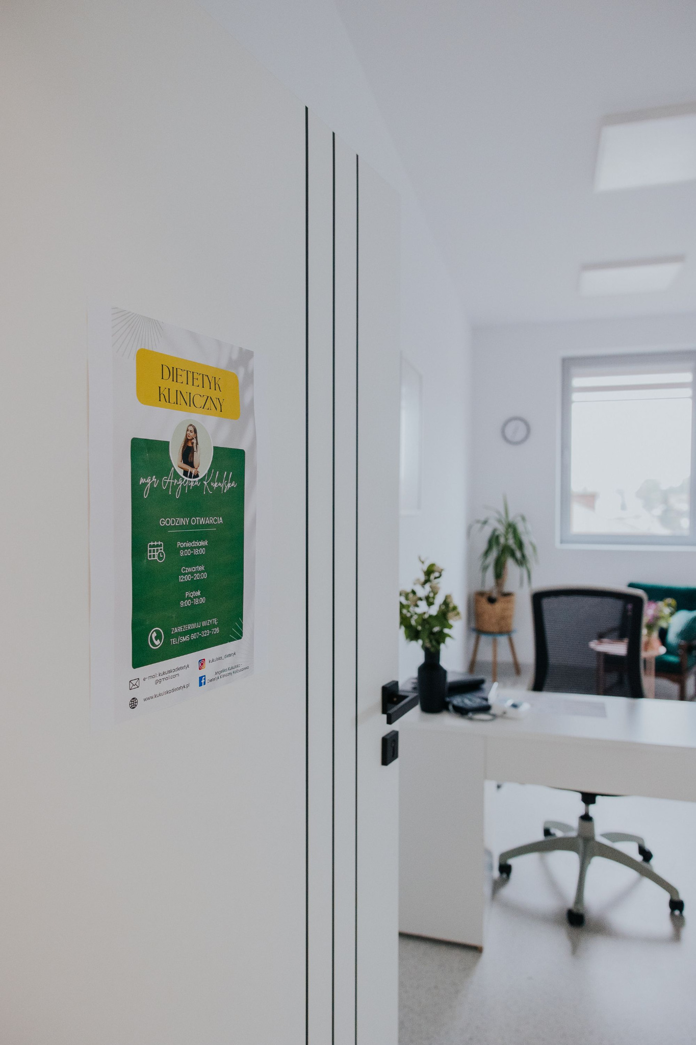 Plakat informacyjny z danymi kontaktowymi i godzinami otwarcia gabinetu na białych drzwiach, w tle jasne wnętrze poczekalni z biurkiem, kwiatami i zielonym fotelem.