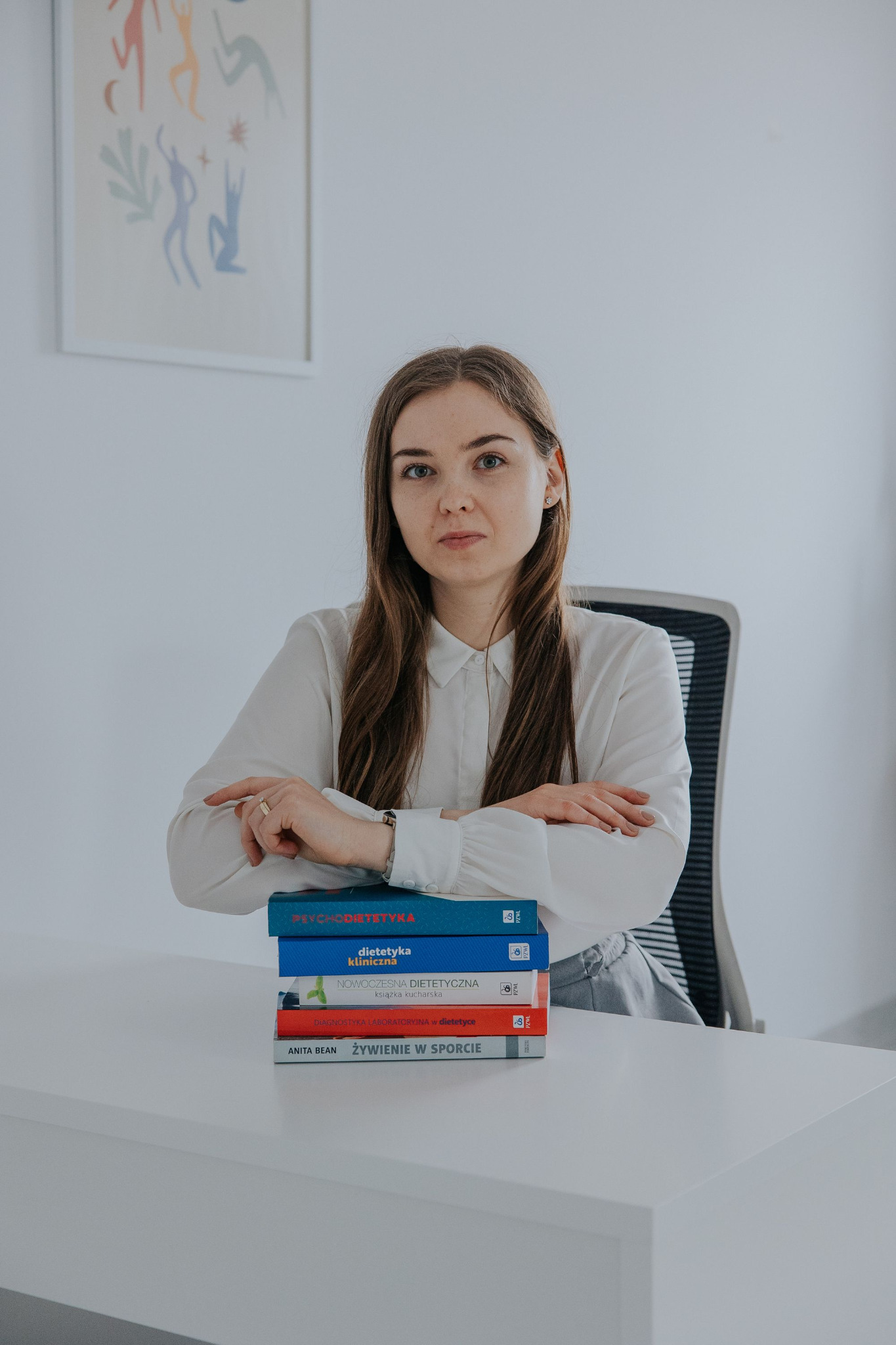 Kobieta w białej koszuli siedzi za białym biurkiem, opierając ręce na stosie książek o tematyce żywienia i dietetyki, z abstrakcyjnym obrazem na ścianie w tle.