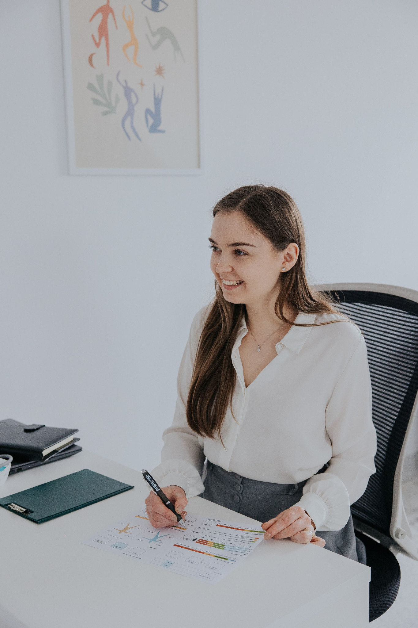 Uśmiechnięta kobieta w białej bluzce analizuje kolorowy wykres na biurku, obok notatnik i klipboard, w tle abstrakcyjny plakat.
