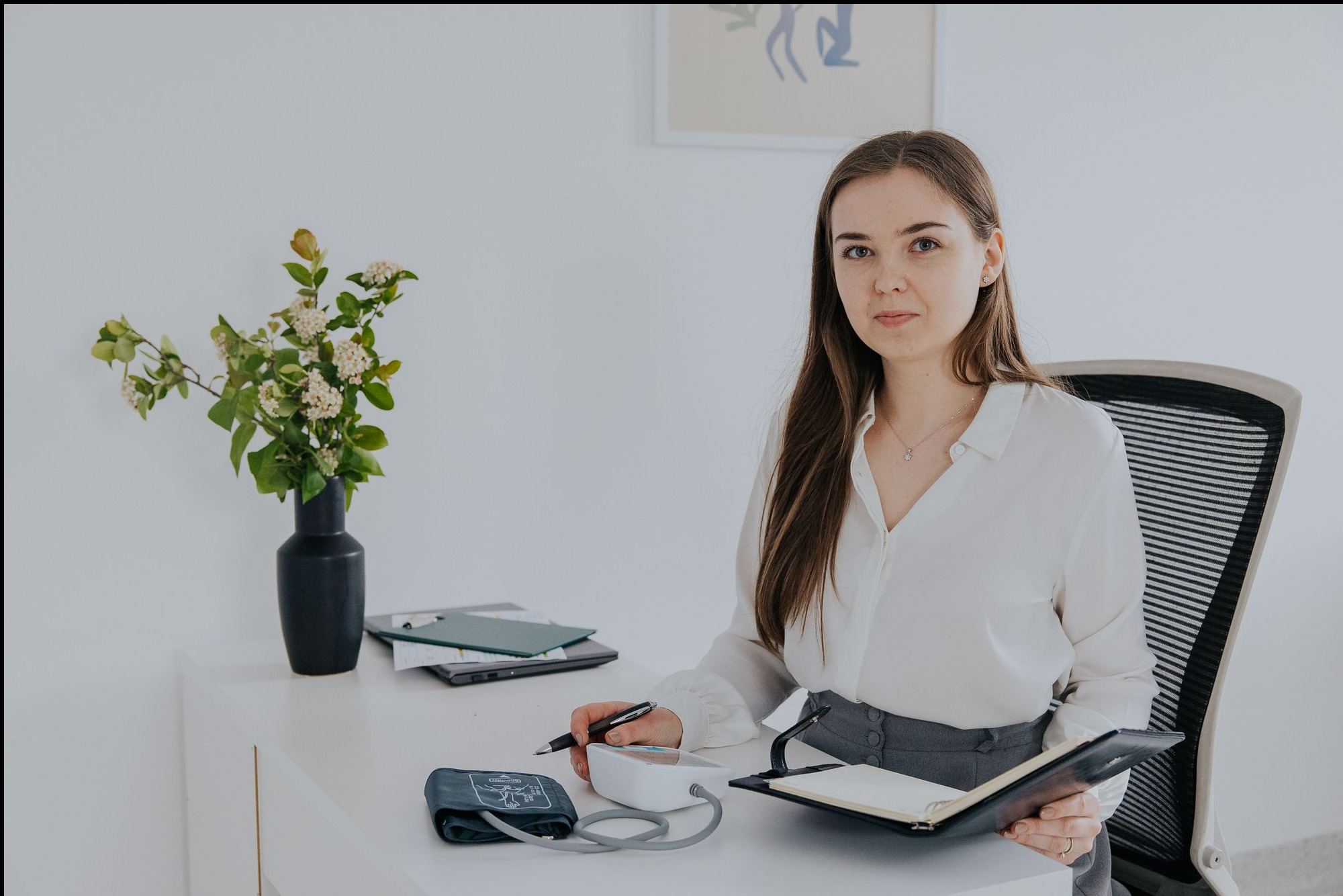 Dietetyczka w białej koszuli, mierząca ciśnienie pacjentowi przy biurku z wazonem kwiatów, notując wyniki w otwartym notatniku.