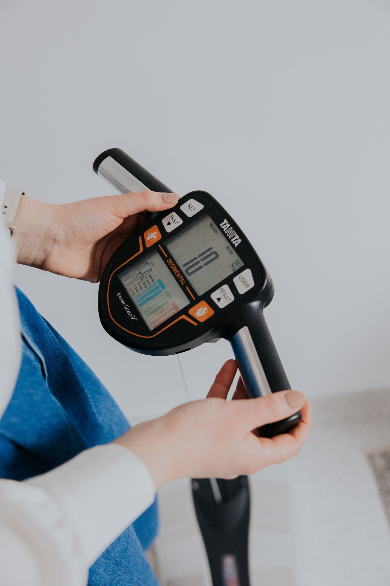 Osoba trzyma w dłoniach analizator składu ciała Tanita InnerScan V z widocznym ekranem i segmentową analizą ciała.