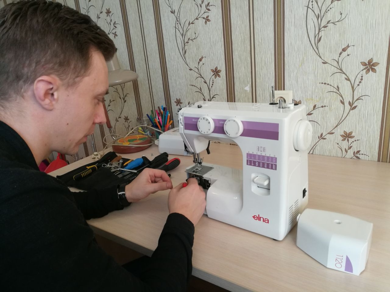 Mężczyzna w czarnym swetrze naprawia białą maszynę do szycia Elna na biurku z zestawem narzędzi, widoczny wzorzysty tapeta w tle.