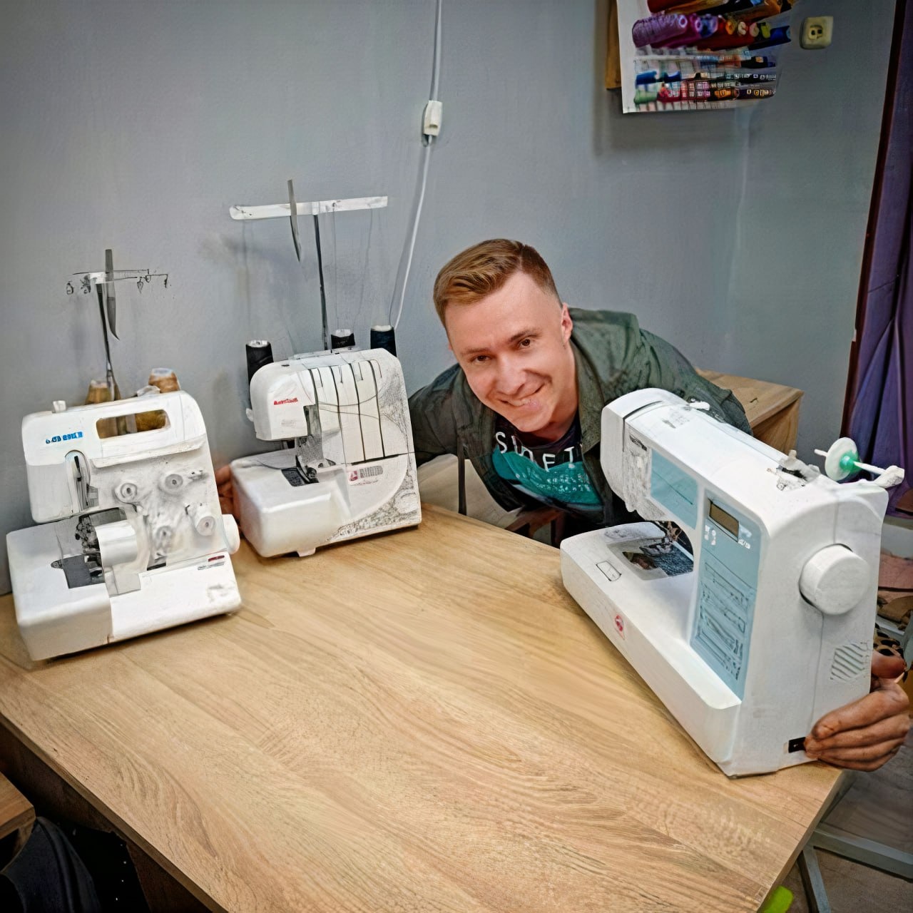 Uśmiechnięty mężczyzna prezentuje trzy białe maszyny do szycia, w tym dwa overloki i jedna maszyna wieloczynnościowa, ustawione na jasnym drewnianym stole w pracowni krawieckiej.