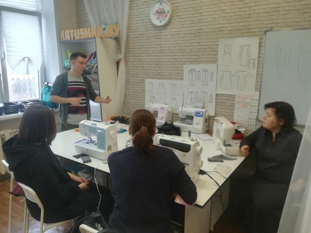 Szkolenie z obsługi maszyn do szycia: instruktor objaśnia działanie maszyny grupie kursantek w pracowni z rysunkami projektów odzieży na ścianie.