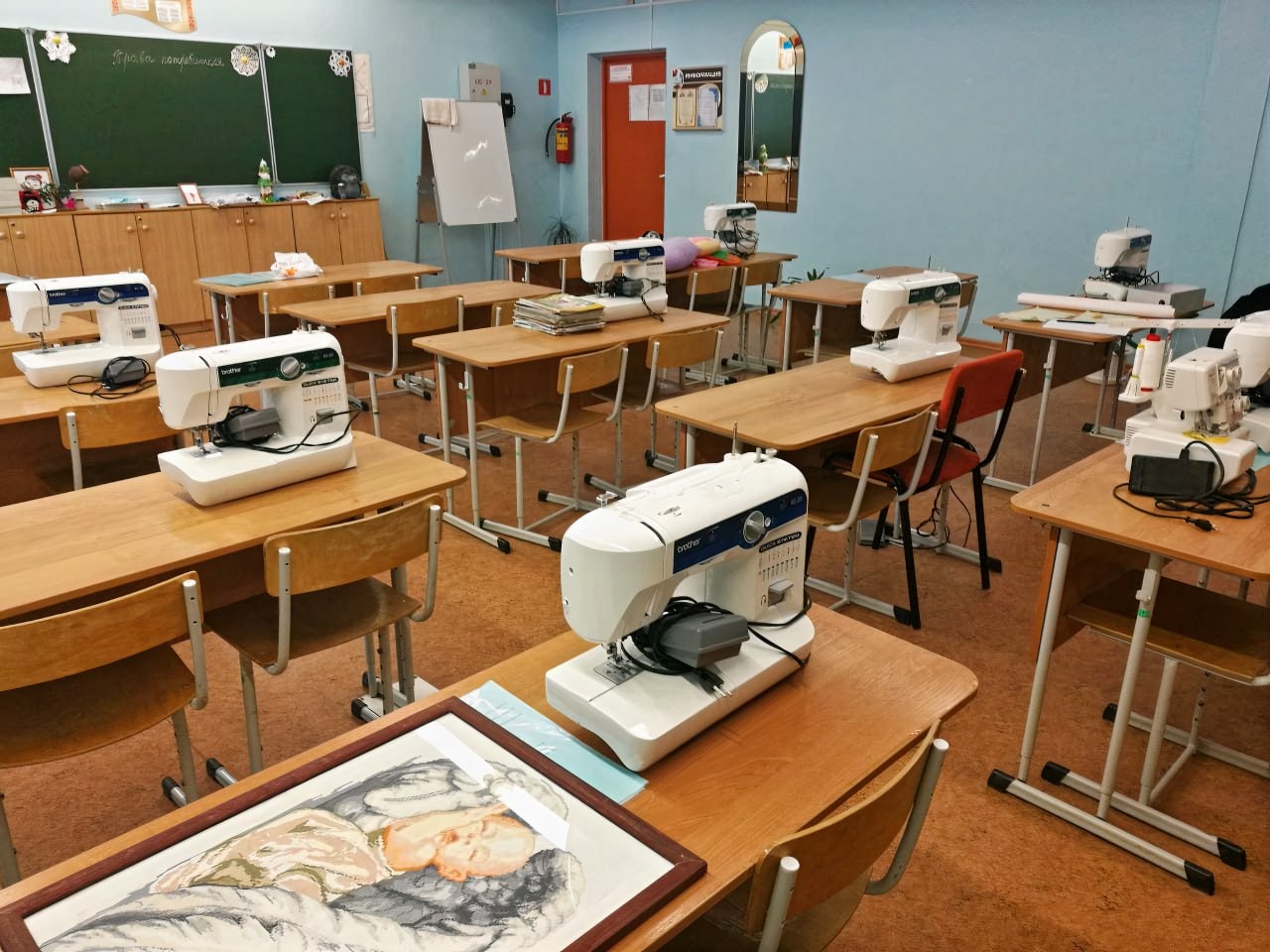 Sala z drewnianymi ławkami, na których stoją białe maszyny do szycia Brother, na pierwszym planie oprawiony obraz z motywem śpiącego dziecka.