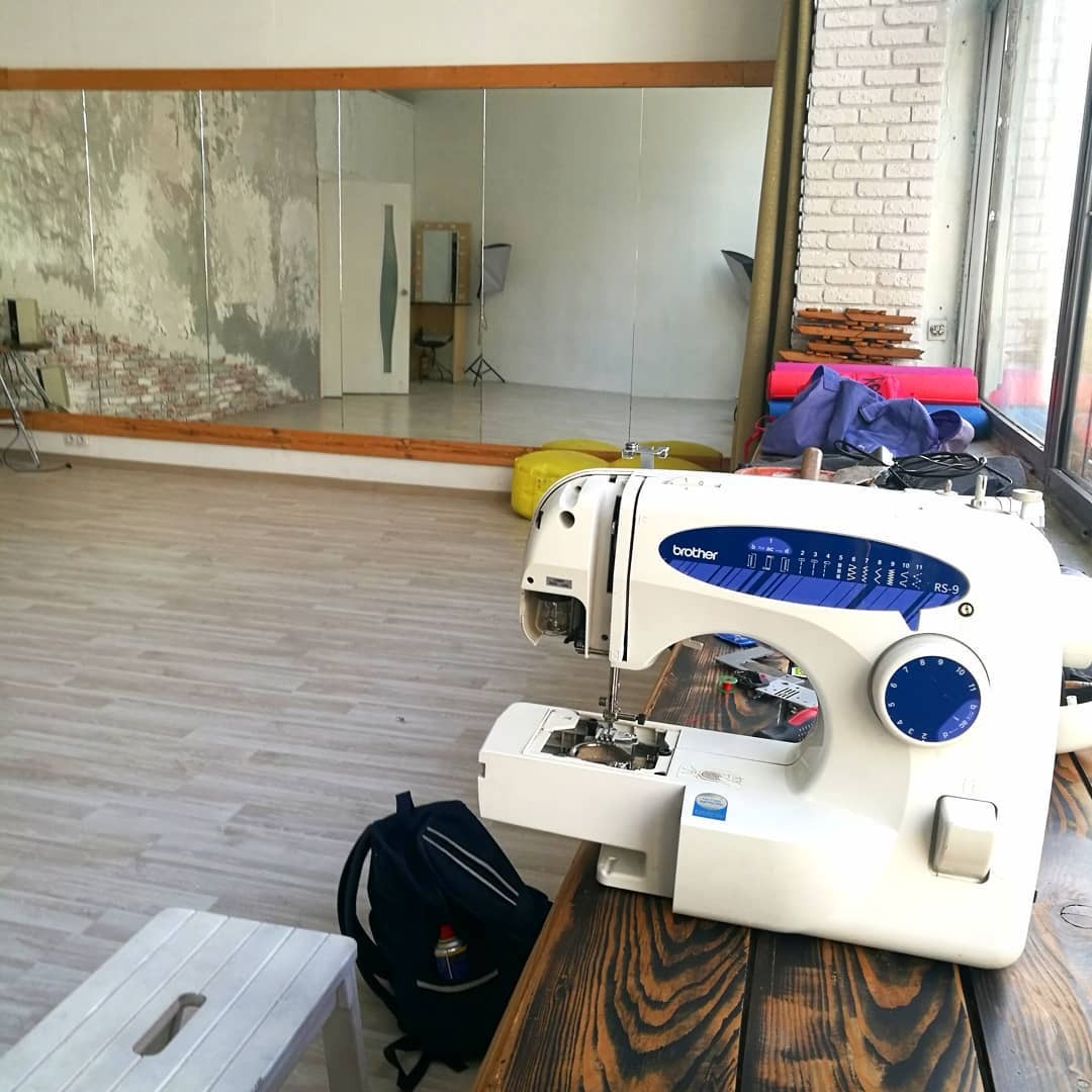 Biała maszyna do szycia Brother RS-9 na drewnianym stole w jasnym pomieszczeniu z dużym lustrem, obok plecak i mały stołek.