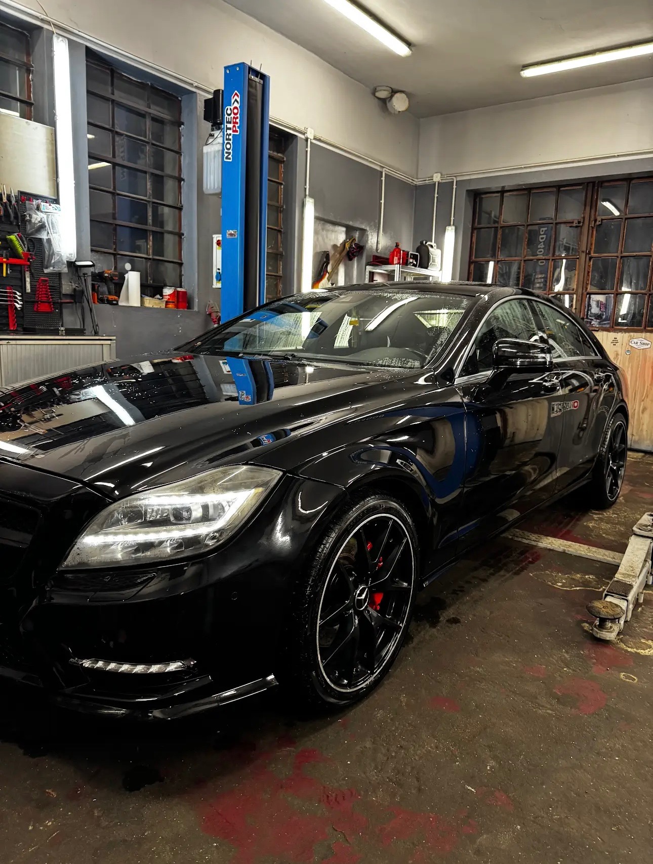 Czarny Mercedes-Benz CLS na podnośniku samochodowym w warsztacie, widoczne narzędzia i wyposażenie warsztatowe w tle.
