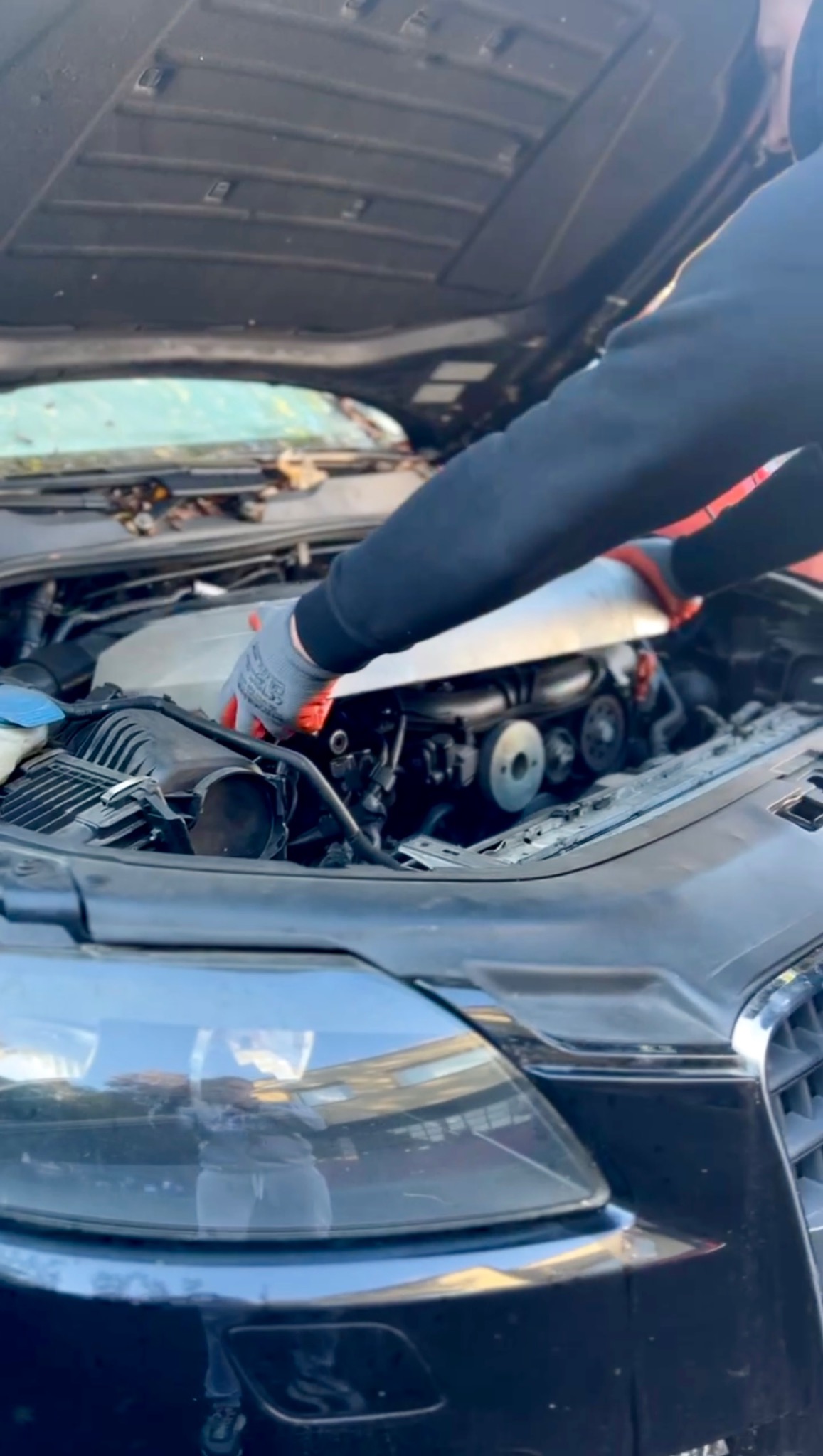 Mechanik w rękawicach roboczych sprawdza silnik samochodu z otwartą maską, widoczne elementy silnika i przednia część pojazdu.