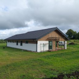 Prywatna - Parterowy dom z szarą dachówką i częściowo otynkowaną białą elewacją, otoczony zielonym trawnikiem, z widocznym rusztowaniem przy ścianie szczytowej, sugerującym trwające prace wykończeniowe elewacji.