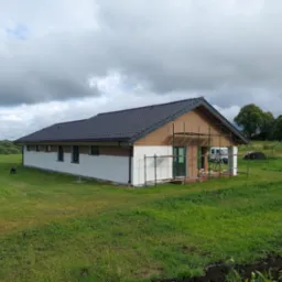 Parterowy dom z szarą dachówką i częściowo otynkowaną białą elewacją, otoczony zielonym trawnikiem, z widocznym rusztowaniem przy ścianie szczytowej, sugerującym trwające prace wykończeniowe elewacji.