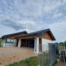 Prywatna - Nowoczesny dom jednorodzinny z białymi ścianami i brązową drewnianą elewacją, widok z zewnątrz na podwórze z żwirową nawierzchnią, częściowo widoczne rusztowanie przy oknie i pies w otwartym garażu.