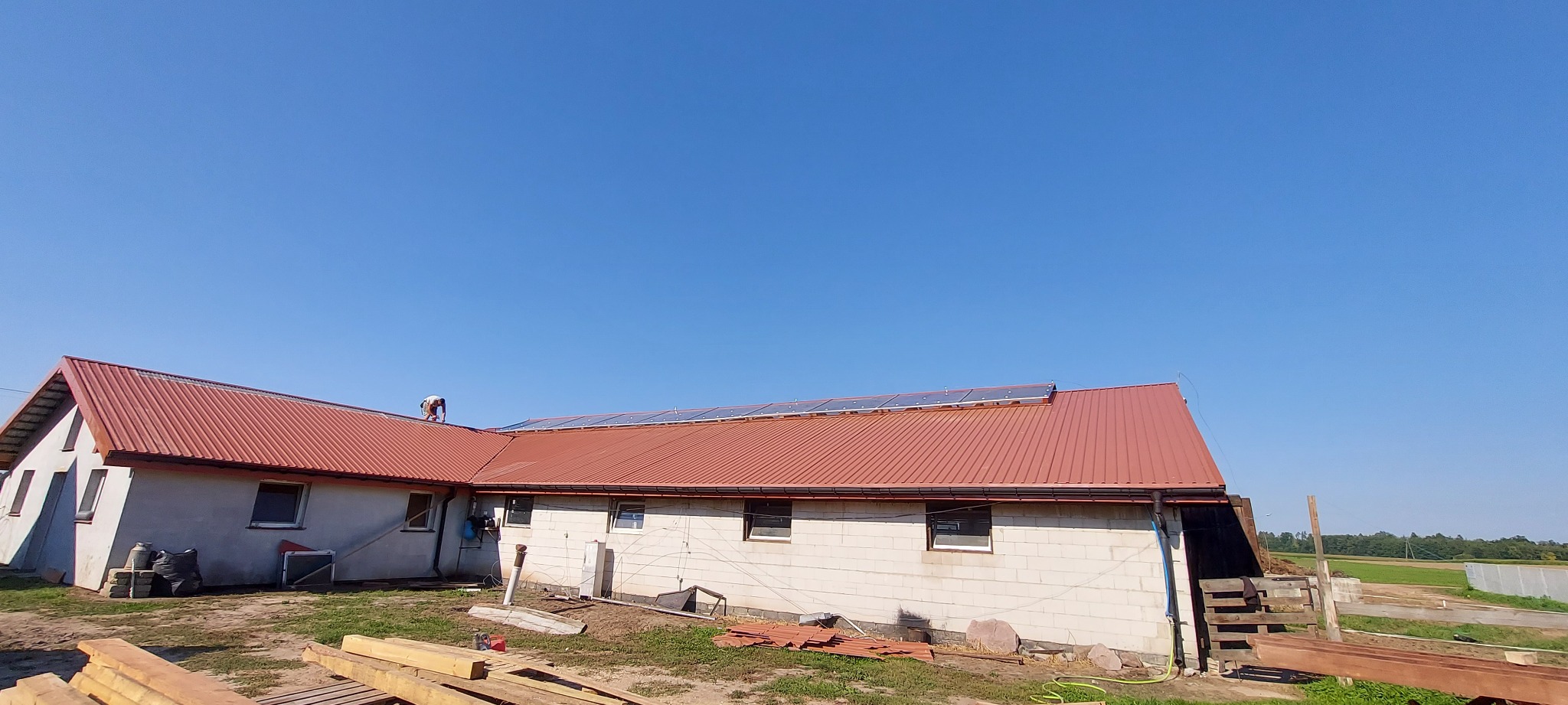 Montaż paneli fotowoltaicznych na dachu domu z blachy trapezowej w słoneczny dzień, widoczny pracownik na dachu i drewniane belki na ziemi.