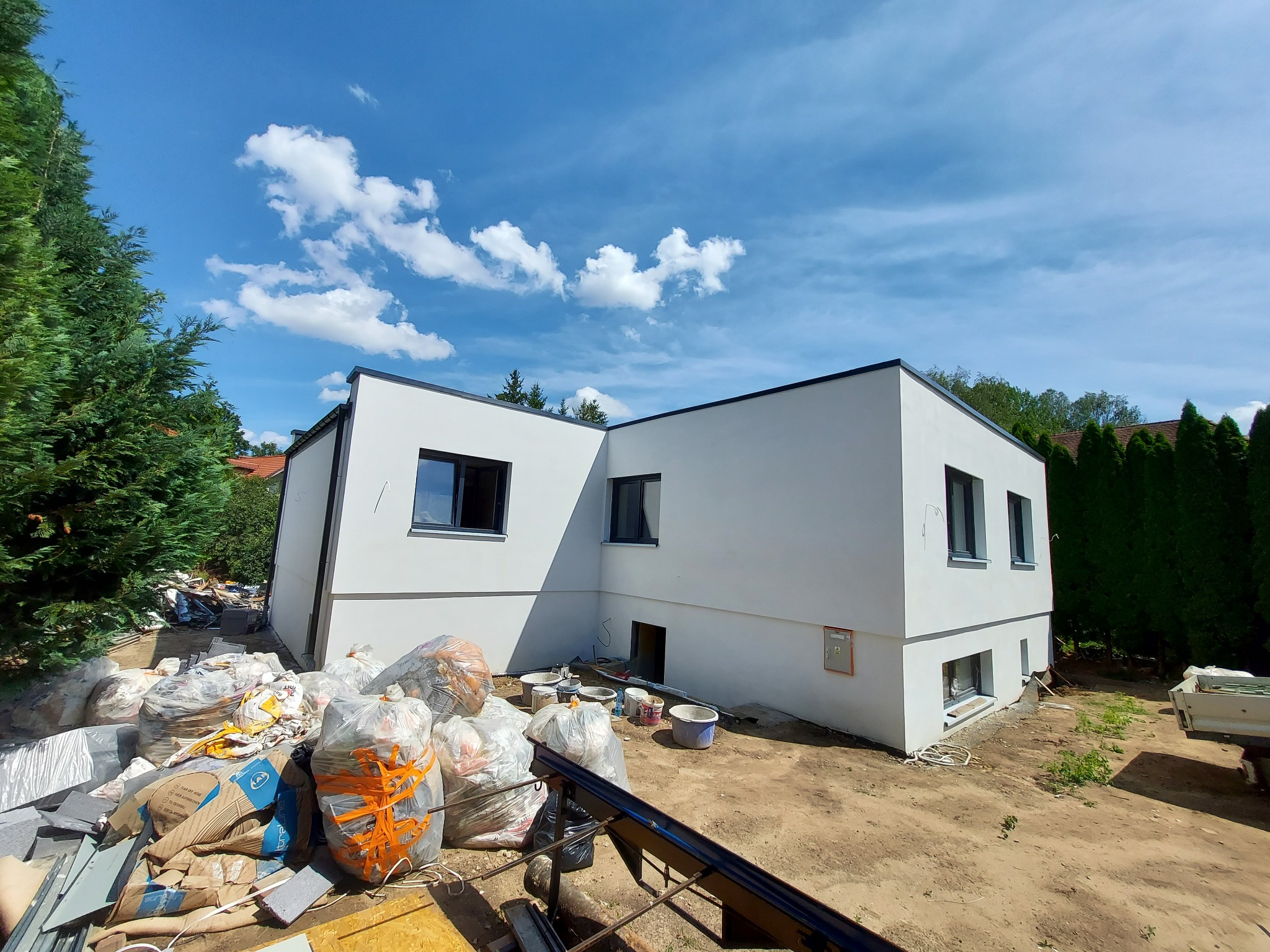 Nowoczesny, dwukondygnacyjny dom z białą elewacją i ciemnymi oknami w trakcie prac wykończeniowych, widoczne worki z materiałami budowlanymi i narzędzia na posesji.