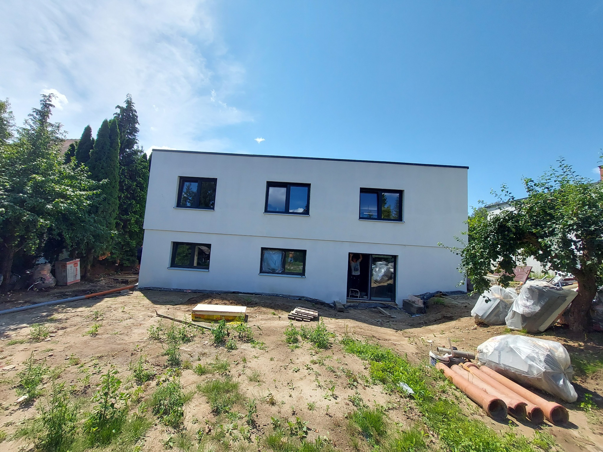 Nowoczesny, dwukondygnacyjny dom z płaskim dachem w trakcie budowy, z widocznymi oknami z czarnymi ramami, otoczony zielenią i materiałami budowlanymi.