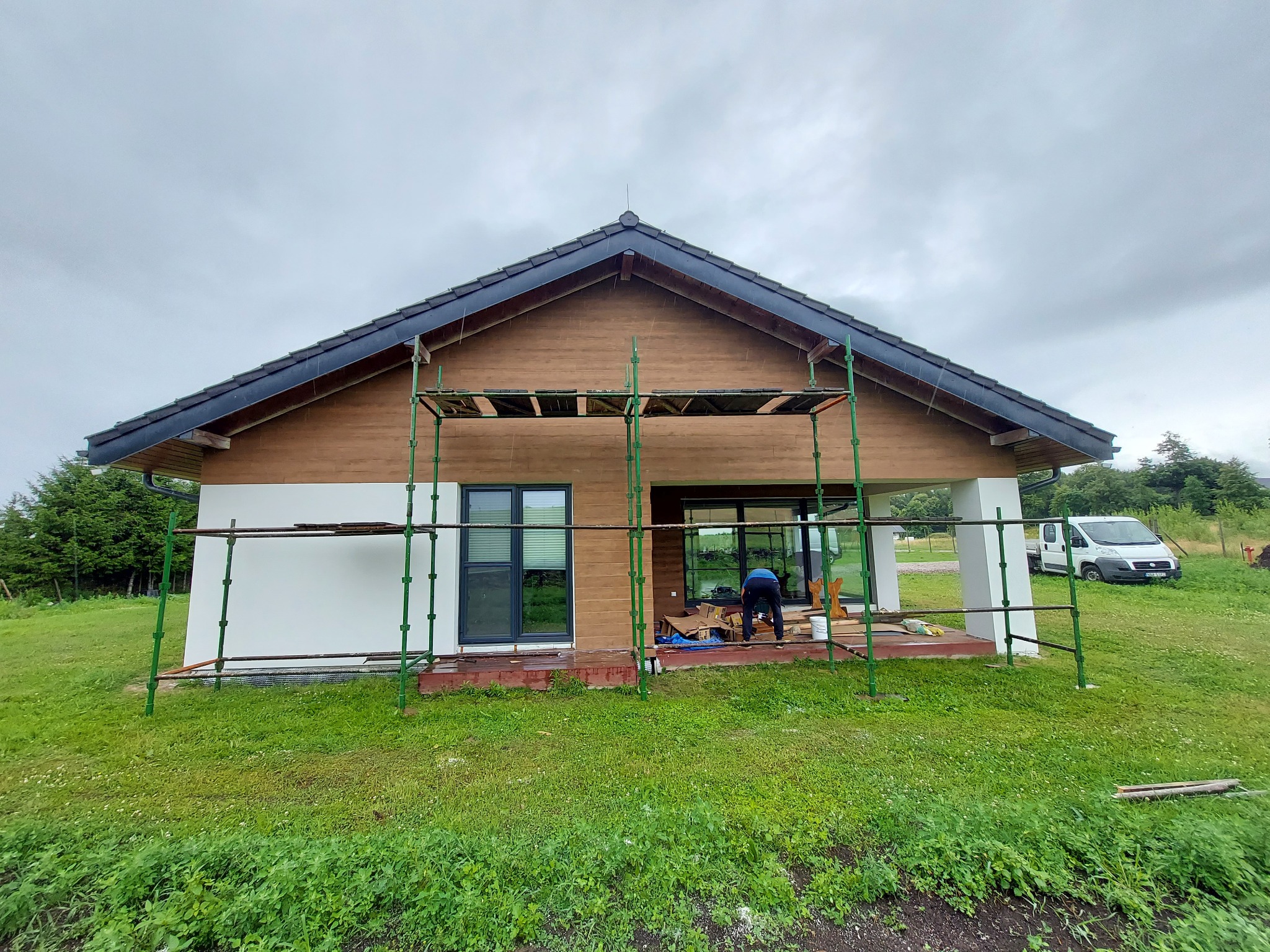 Dom jednorodzinny w trakcie prac elewacyjnych, widoczne rusztowania, częściowo ocieplona ściana, drewniana fasada i pracownik przy oknie.