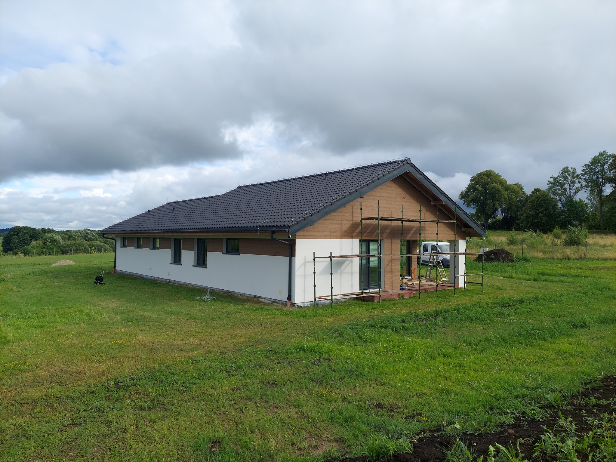 Parterowy dom z szarą dachówką i częściowo otynkowaną białą elewacją, otoczony zielonym trawnikiem, z widocznym rusztowaniem przy ścianie szczytowej, sugerującym trwające prace wykończeniowe elewacji.