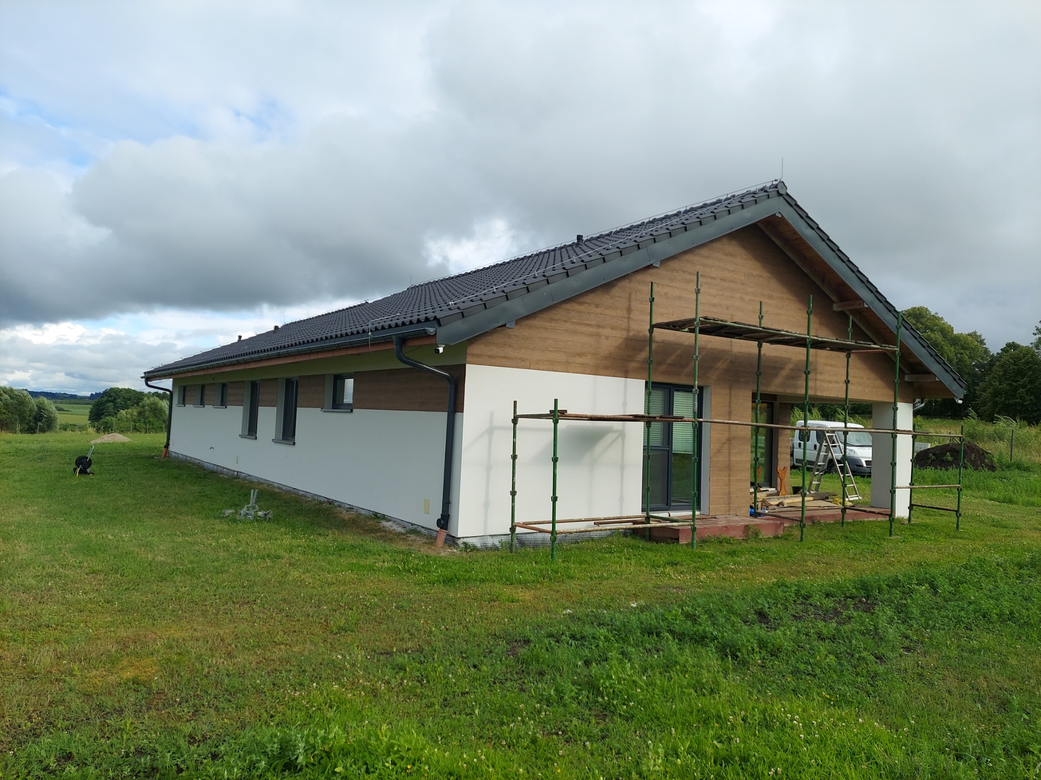 Parterowy dom jednorodzinny z drewnianą elewacją na wysokości dachu, częściowo ocieplony, z rozstawionym rusztowaniem i widocznymi elementami wykończeniowymi.