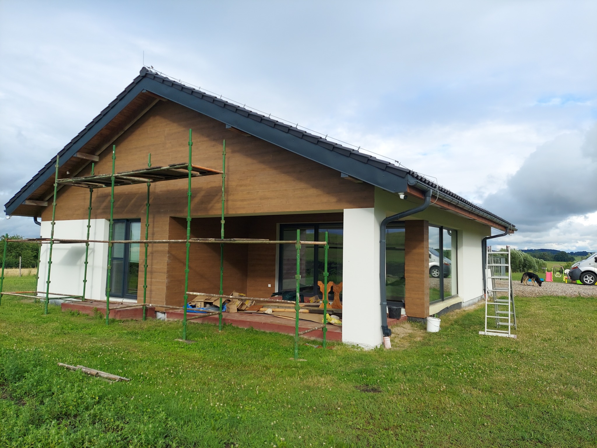 Nowoczesny dom jednorodzinny z drewnianą elewacją, częściowo ocieplony, z widocznymi rusztowaniami i drabiną, sugerującymi trwające prace wykończeniowe na zewnątrz budynku, na tle zielonego...