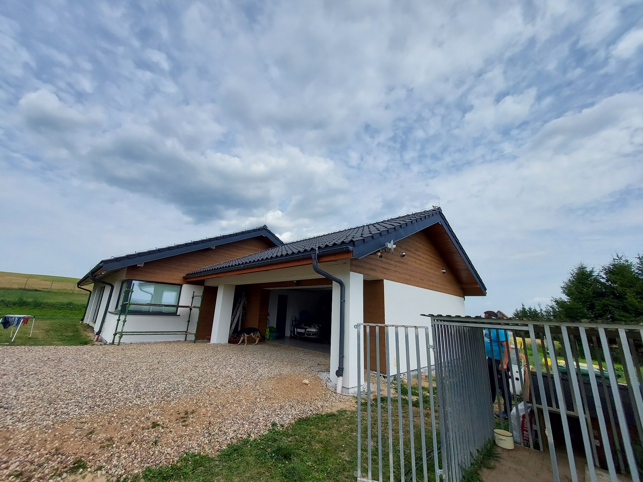 Nowoczesny dom jednorodzinny z białymi ścianami i brązową drewnianą elewacją, widok z zewnątrz na podwórze z żwirową nawierzchnią, częściowo widoczne rusztowanie przy oknie i pies w otwartym garażu.