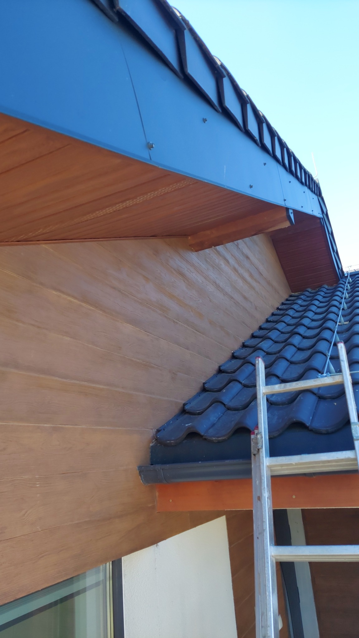 Fragment domu z ciemną dachówką, drewnopodobną elewacją i metalową drabiną opartą o dach, widok z dołu.
