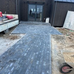 Konrad Szczepański - Ułożona ścieżka z szarej kostki brukowej prowadząca do nowoczesnego domu z ciemnego drewna; widoczny wózek z piaskiem i przyczepa z narzędziami.