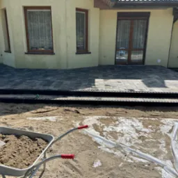 Ułożona kostka brukowa przed domem z widocznym wózkiem wypełnionym piaskiem i łopatą, przygotowanie terenu pod dalsze prace brukarskie.