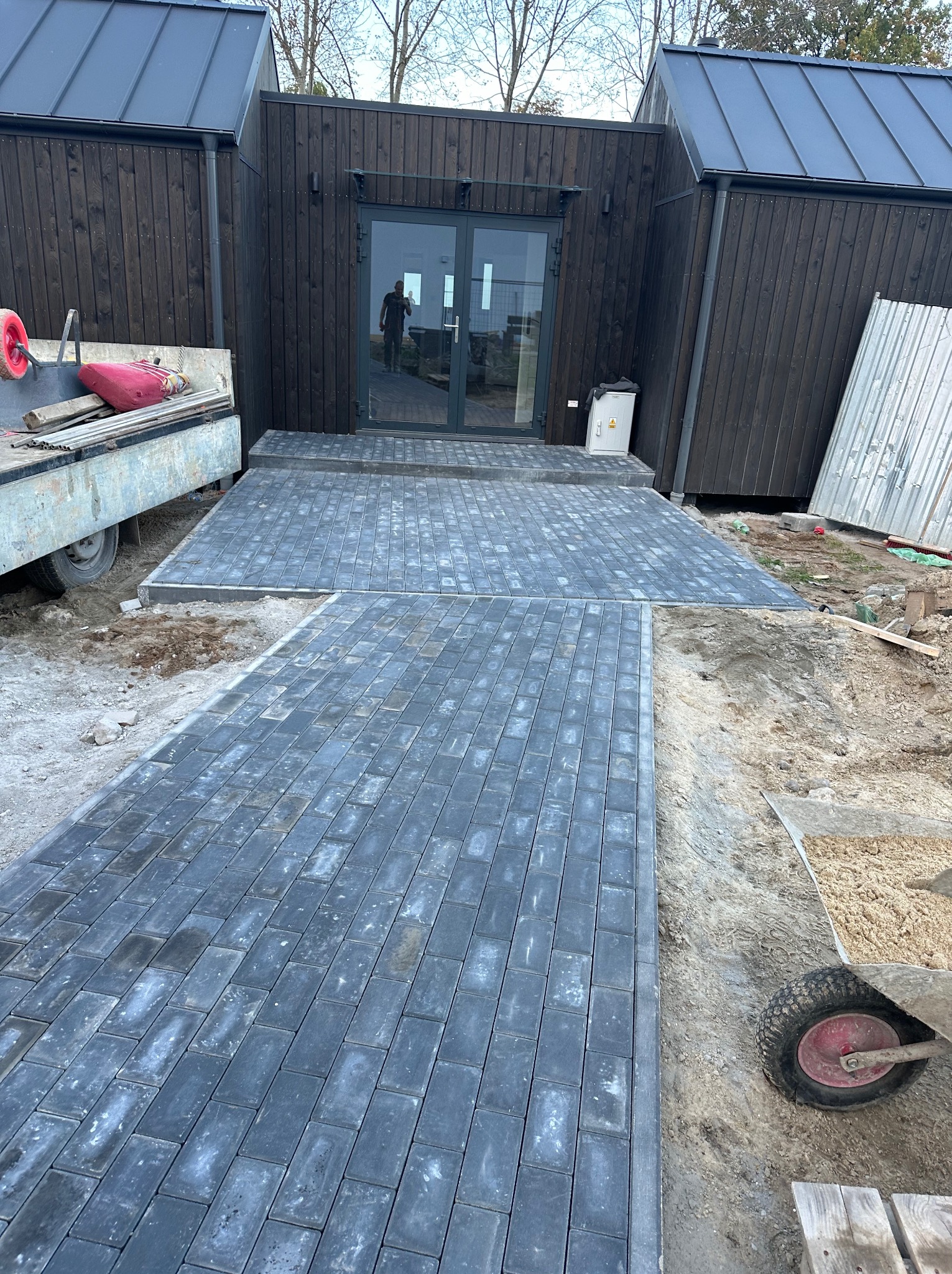 Ułożona ścieżka z szarej kostki brukowej prowadząca do nowoczesnego domu z ciemnego drewna; widoczny wózek z piaskiem i przyczepa z narzędziami.