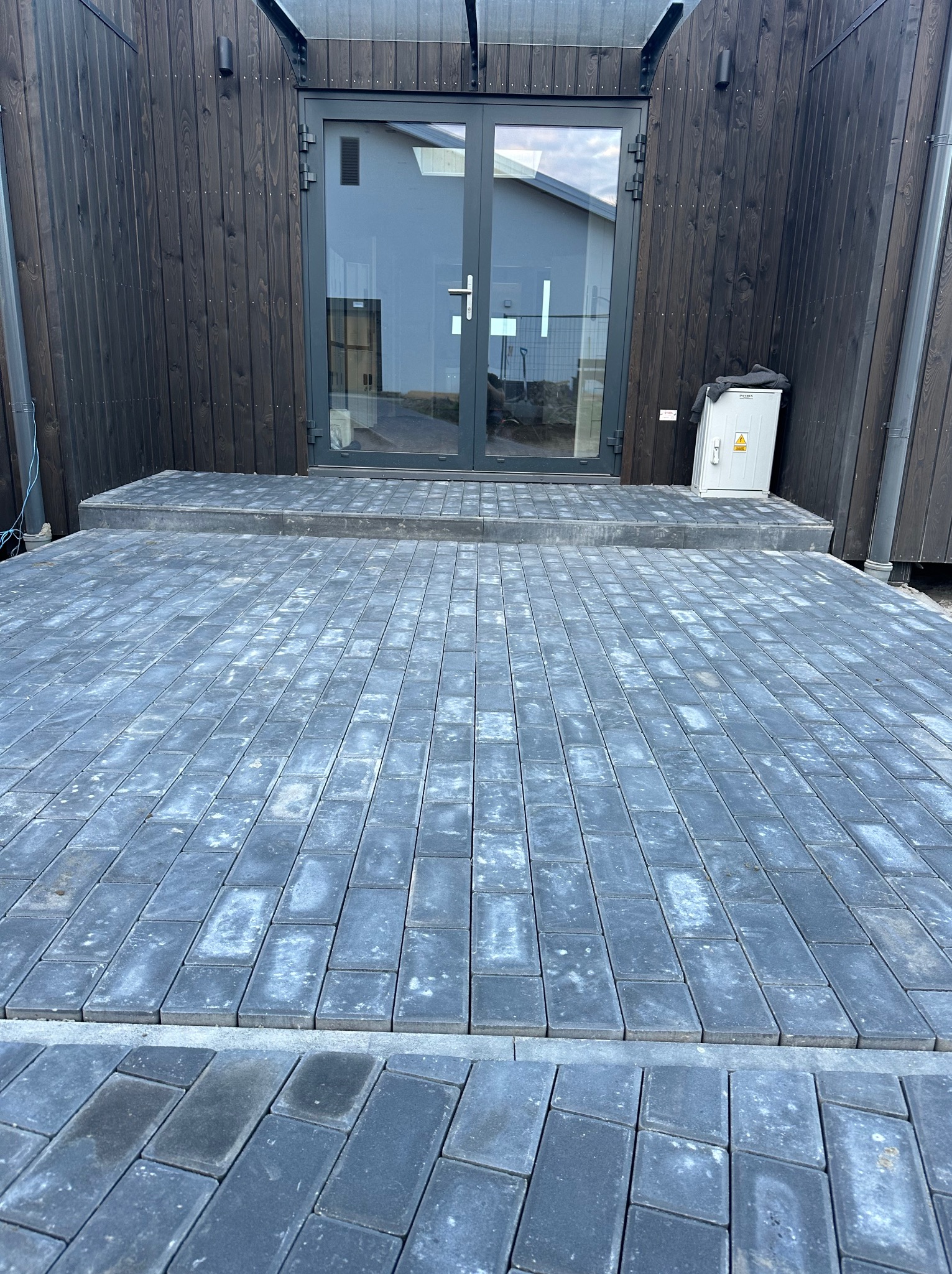 Ułożona szara kostka brukowa prowadząca do wejścia z drzwiami do budynku o ciemnej, drewnianej elewacji. Widoczna skrzynka elektryczna na ścianie.