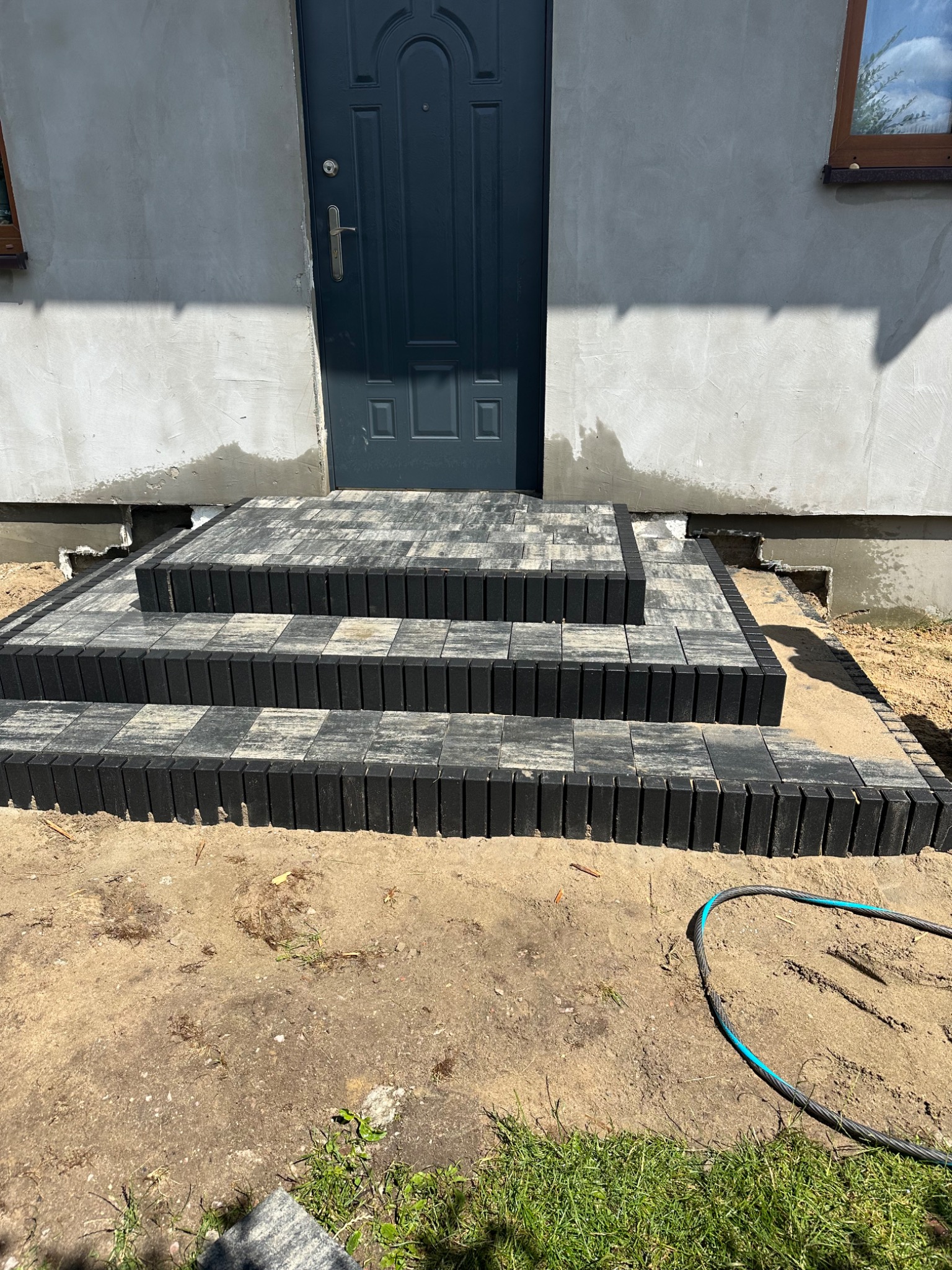 Schody wejściowe do domu wyłożone szaro-czarną kostką brukową, z widocznym fragmentem ściany budynku i niebieskiego węża ogrodowego leżącego na ziemi.