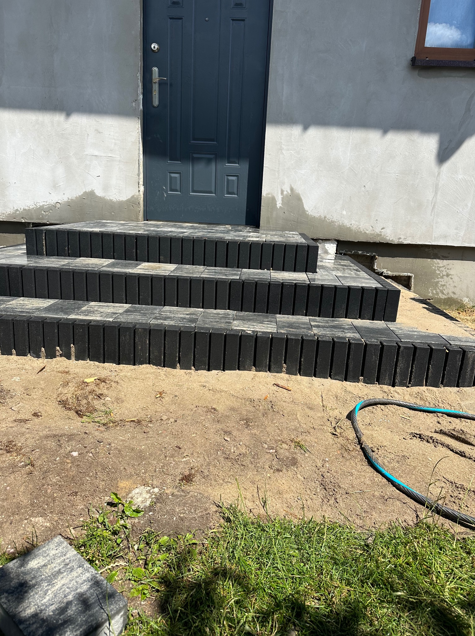 Nowe schody wejściowe z kostki brukowej w odcieniach szarości, prowadzące do ciemnych drzwi wejściowych, widoczne niedokończone prace ziemne i fragment węża ogrodowego.