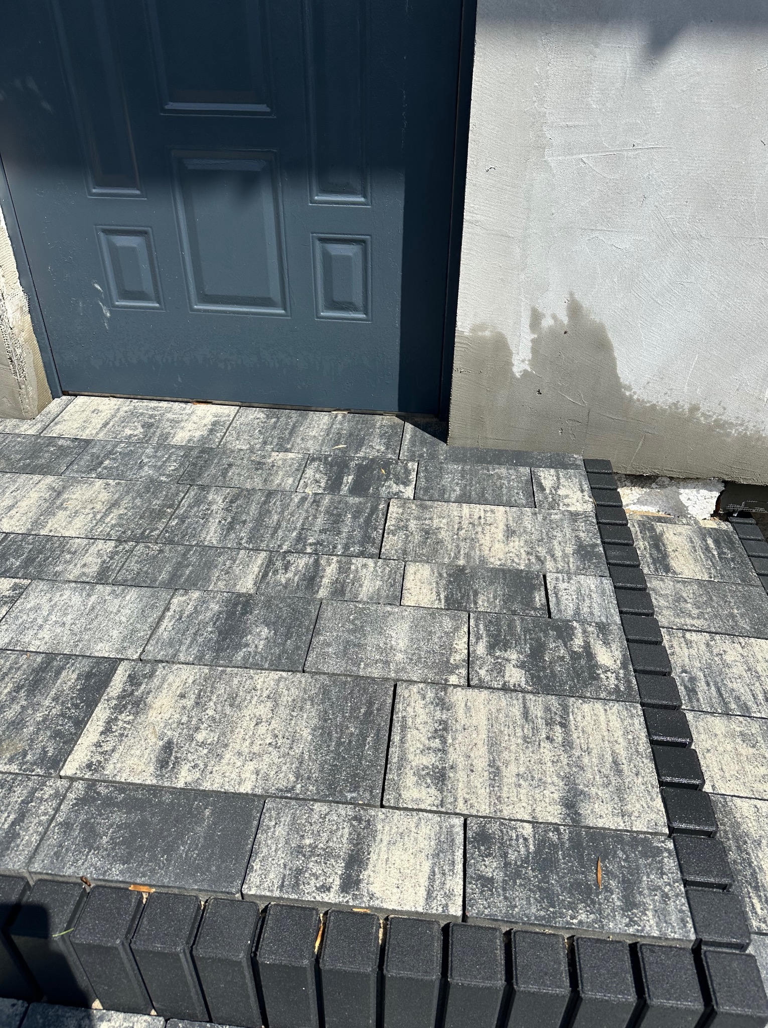Ułożona kostka brukowa w odcieniach szarości i bieli prowadząca do ciemnoszarych drzwi wejściowych, z widocznym fragmentem otynkowanej ściany po prawej stronie.