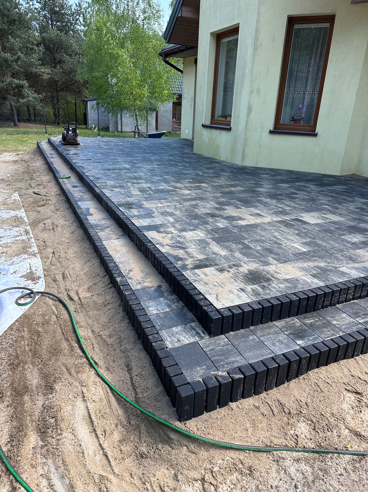 Ułożona kostka brukowa w odcieniach szarości tworząca taras przy budynku, z widocznymi obrzeżami z ciemnej kostki i ubijarką w tle.