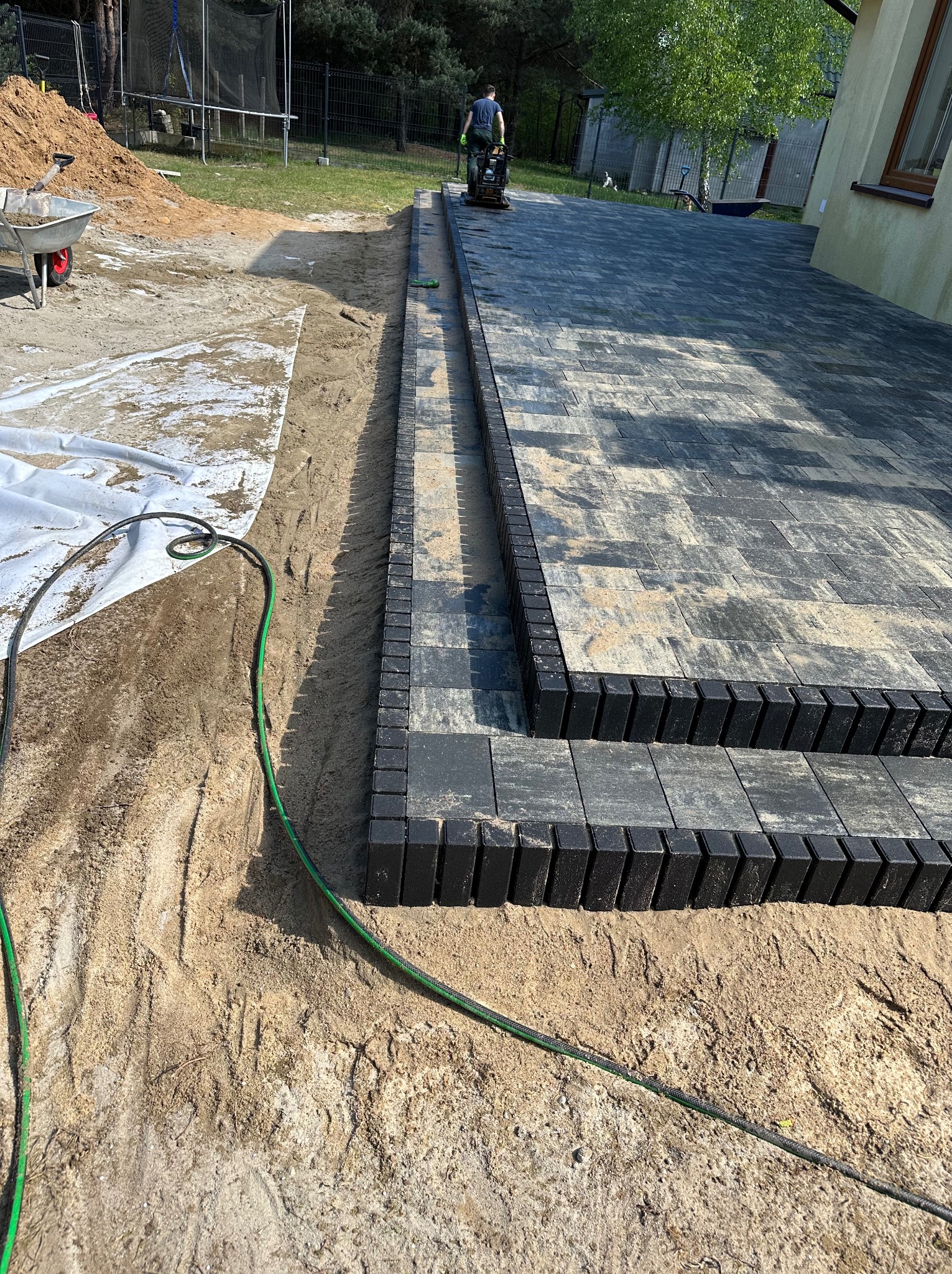 Częściowo ułożona nawierzchnia z kostki brukowej w odcieniach szarości i czerni, widoczny pracownik zagęszczający teren, obok przygotowany teren z piaskiem, w tle drzewa i budynek.