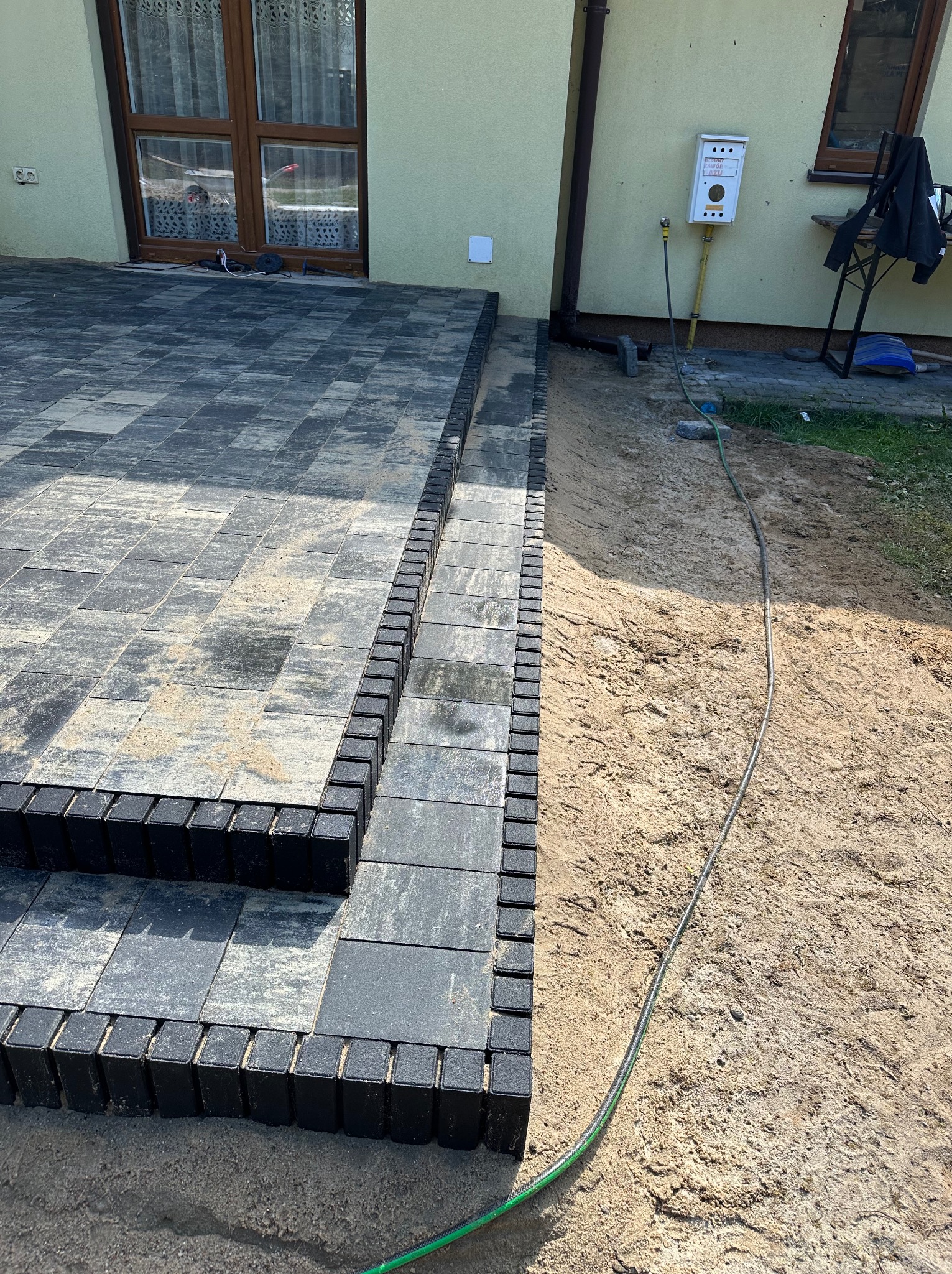 Ułożona kostka brukowa w odcieniach szarości tworząca schody przed budynkiem, obrzeża z ciemnej kostki, widoczny fragment trawnika i piasku.