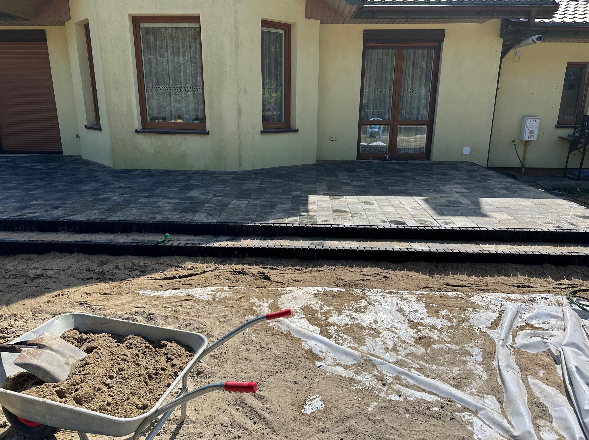 Ułożona kostka brukowa przed domem z widocznym wózkiem wypełnionym piaskiem i łopatą, przygotowanie terenu pod dalsze prace brukarskie.