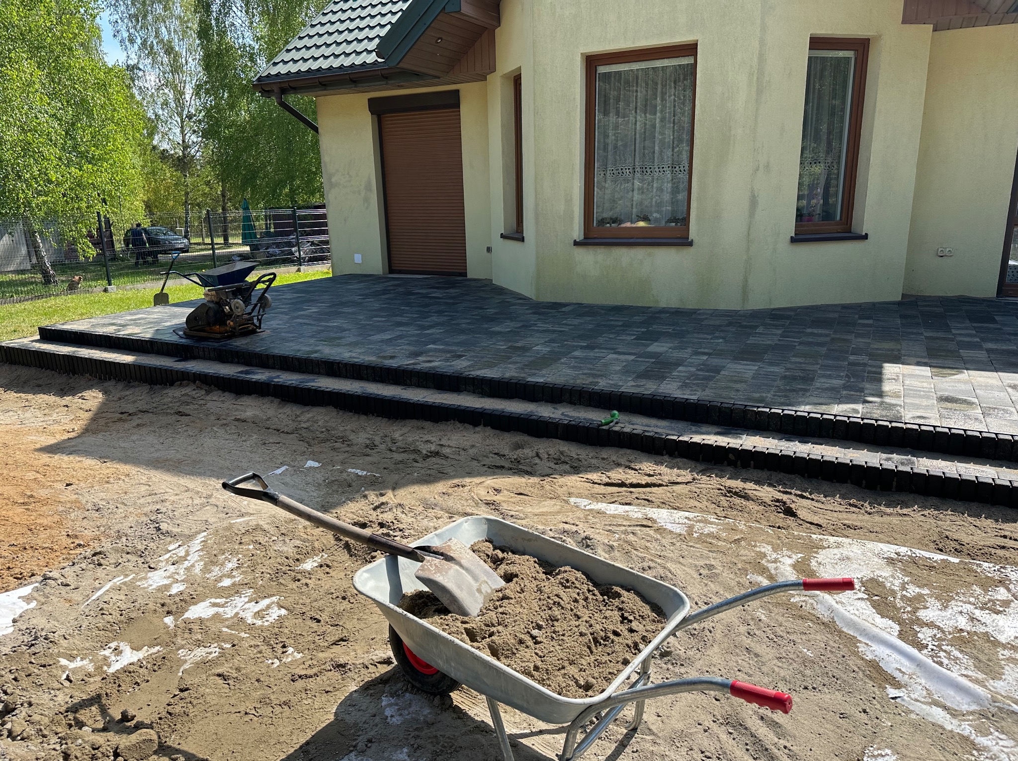 Taczka z piaskiem i łopatą na tle świeżo ułożonej kostki brukowej przed domem, widoczny ubijak wibracyjny i fragment elewacji.