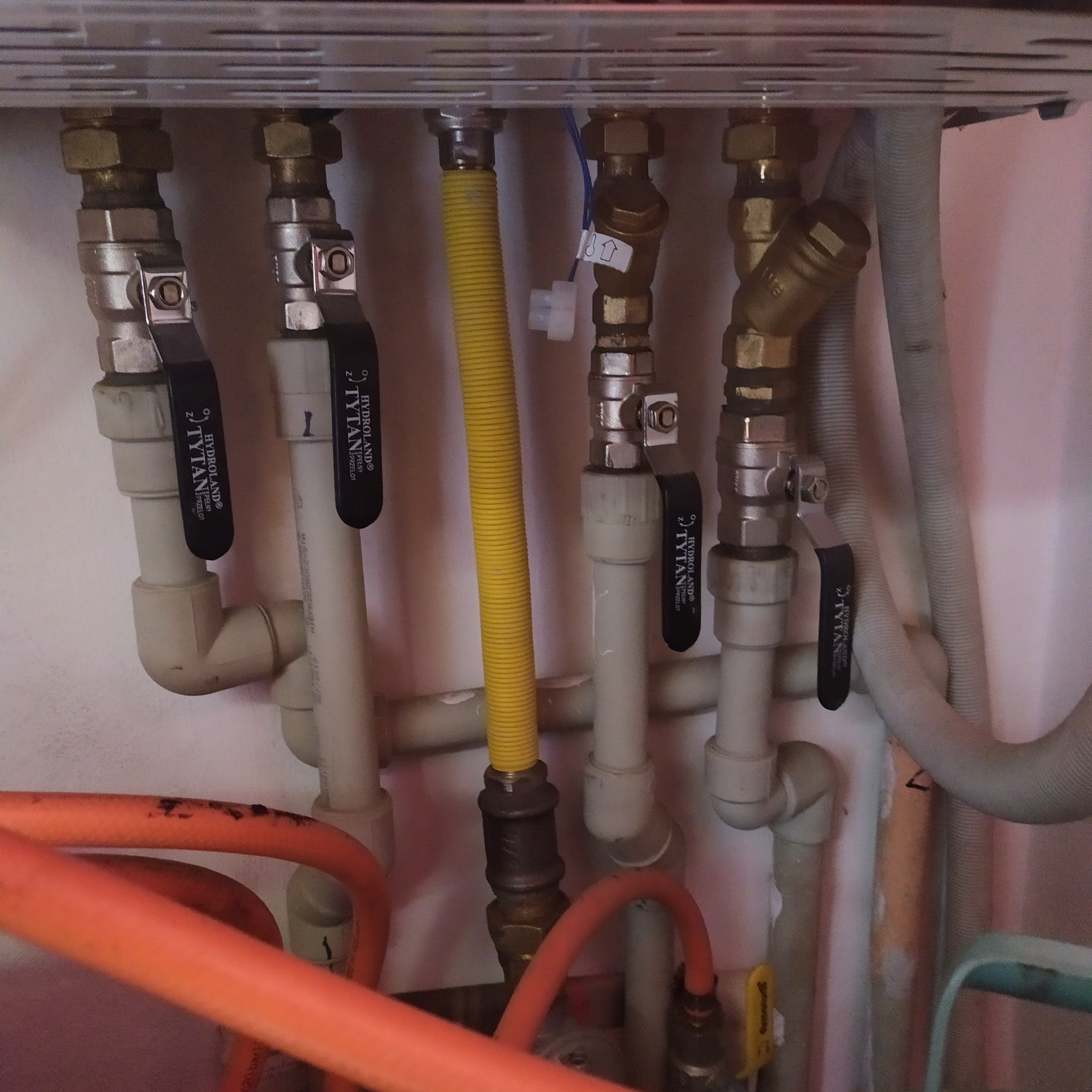 Kompleksowy układ rur z zaworami marki Tytan, z żółtą rurą gazową w centrum, tworzący instalację hydrauliczną pod blatem. Widoczne złącza i elementy mocujące.