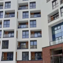 Nowoczesny budynek mieszkalny z ciemnymi oknami i balkonami z przezroczystymi balustradami, fasada w odcieniach bieli i szarości, częściowo wykończona cegłą.